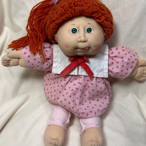 May include: A vintage Cabbage Patch Kid doll with a red yarn hairstyle and green eyes. The doll wears a pink dress with heart patterns, a white collar, and a red bow. It also has pink leggings and fabric arms and legs.