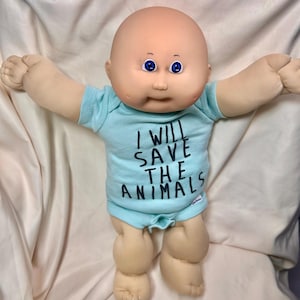 May include: A Cabbage Patch Kid doll with a bald head and blue eyes, wearing a light blue onesie. The onesie has the text "I WILL SAVE THE ANIMALS" printed on the front. The doll's arms are outstretched.