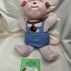 May include: A Cabbage Patch Kids plush doll with pink skin, white hair, and blue denim overalls. The doll has a brown collar with a pendant and is holding a green registration certificate.