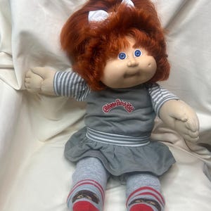 May include: A Cabbage Patch Kids doll with bright red hair, blue eyes, and a dimpled face. The doll wears a gray dress with white striped sleeves, a white bow in its hair, and gray leggings featuring red and white striped cuffs with small graphic prints.