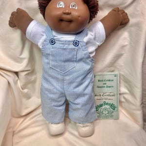 May include: A Cabbage Patch Kids doll with brown skin, brown hair, and open arms. The doll wears a white shirt under blue and white striped overalls. A birth certificate and adoption papers are visible.