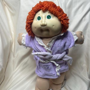 May include: A Cabbage Patch Kids doll with red yarn hair and green eyes. The doll is wearing a light purple terry cloth robe with white lace trim and a matching belt. The doll's arms are outstretched.