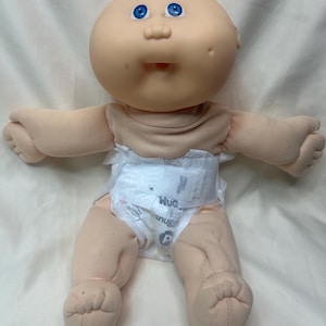 May include: A doll with a round, light-toned vinyl head, bright blue eyes, and a soft fabric body. The doll has a dimple on the left cheek and is wearing a white disposable diaper with the text Huggies Snug & Dry printed on the front panel.