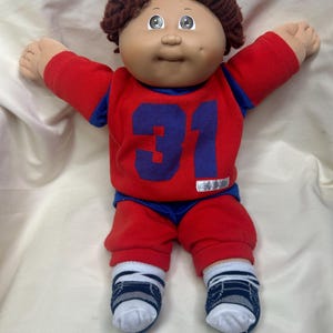 May include: A vintage Cabbage Patch Kid doll with brown yarn hair, a fair complexion, and blue eyes. The doll is wearing a red and blue jersey with the number 31, red pants, and blue and white socks. The doll's arms are outstretched.