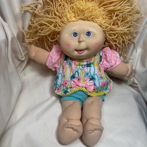 May include: A vintage Cabbage Patch Kid doll with blonde yarn hair, blue eyes, and rosy cheeks. The doll wears a pink and white floral print top, blue shorts, and has a yellow trim. The doll's arms and legs are made of a soft material.