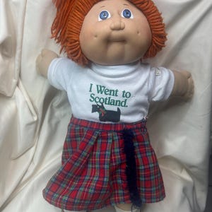 May include: A vintage doll with red yarn hair, blue eyes, and a white t-shirt that says "I Went to Scotland." The doll wears a red and green plaid skirt, white socks, and brown shoes. A gray bow adorns the doll's hair.