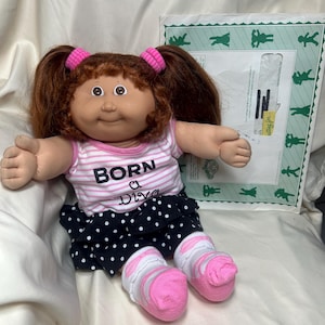 May include: A doll with brown hair in pigtails, wearing a pink and white striped shirt with the text BORN a Diva, a black skirt with white polka dots, and white socks with pink trim. The doll is positioned in front of a white envelope with green graphics.