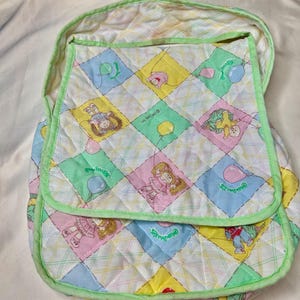 May include: Quilted baby carrier with a colorful pattern featuring cartoon characters, balloons, and the text "Cabbage Patch Kids." The carrier has a light green trim and a flap closure. The background is a light-colored fabric.