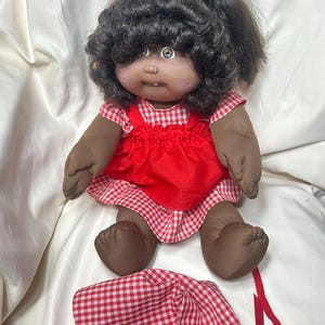 May include: A doll with dark skin, brown eyes, and dark curly hair tied in a ponytail. The doll wears a red and white gingham dress with a solid red apron overlay. A matching red and white gingham bonnet with red ribbon ties is placed in front.