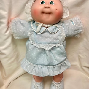 May include: A vintage Cabbage Patch Kid doll. The doll has a round face with rosy cheeks, green eyes, and a small smile. It wears a light blue dress with white polka dots, lace trim, and a matching bonnet. White shoes complete the outfit.