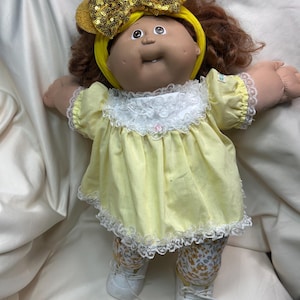 May include: A vintage Cabbage Patch Kid doll with brown hair and a yellow headband with a sequined bow. The doll wears a yellow dress with white lace trim, floral patterned leggings, and white shoes.