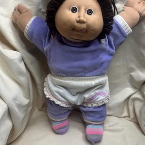 May include: A vintage doll with a soft, vinyl face and brown yarn hair. The doll wears a lavender top and pants, white ruffled shorts, and striped socks. The doll's arms are outstretched.