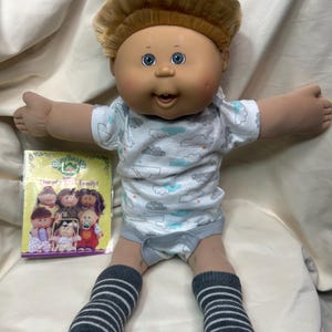 May include: A Cabbage Patch Kids doll with blonde hair, blue eyes, and a smiling expression. The doll wears a white onesie with a cloud pattern, gray shorts, and striped socks. A Cabbage Patch Kids video box is next to the doll.