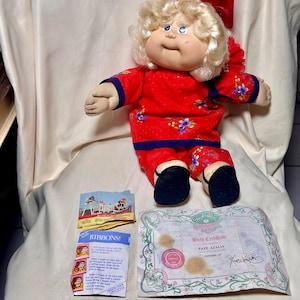 May include: A vintage Cabbage Patch Kids doll, Faye Azalia, wearing a red outfit with blue floral accents and a matching red bow. Includes a birth certificate and a "Ribbons!" instruction sheet.
