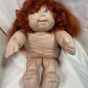 May include: A vintage doll with a soft, flesh-toned body and limbs. The doll has a round face, blue eyes, and a small, open mouth. It has a full head of bright red, curly hair. The doll is a classic toy.