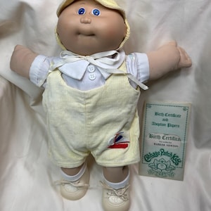May include: A Cabbage Patch Kids doll wearing a yellow corduroy outfit, matching hat, and white shoes. The doll has blue eyes and a birth certificate is included. The outfit has a small sailboat graphic.