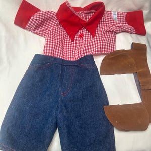 May include: A doll outfit consisting of a short-sleeved shirt with a red and white gingham pattern, a solid red collar, and red sleeve cuffs. The set includes dark blue denim pants with red stitching and a separate brown faux leather belt piece.
