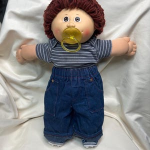May include: A vintage doll with a round face, brown yarn hair, and a pacifier. The doll wears a gray and white striped shirt and denim pants. The doll's arms are outstretched.