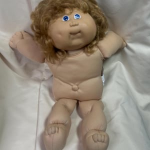 May include: A vintage Cabbage Patch Kid doll with light skin, blue eyes, and light brown curly hair. The doll is nude, with a soft, plush body and detailed facial features. The doll's arms are outstretched.