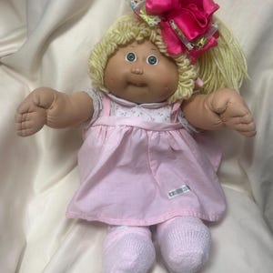 May include: A vintage Cabbage Patch Kids doll with blonde yarn hair and green eyes. The doll wears a pink dress, white socks, and a large pink bow. The doll's arms are outstretched.