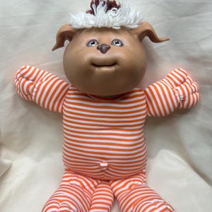 May include: A plush doll with a tan vinyl head, brown nose, and white and brown yarn hair. The doll features a soft body wearing an orange and white horizontally striped long-sleeved onesie with a small orange button at the center of the waist.