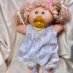 May include: A vintage Cabbage Patch Kid doll with light brown yarn hair styled in pigtails, secured with pink bows and white ribbons. The doll has blue eyes and a yellow pacifier. It wears a white floral print romper with lace trim and white shoes.