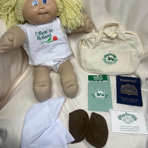 May include: A Cabbage Patch Kids doll with blonde yarn hair, wearing a white t-shirt that says "I Went to Holland." Accessories include a cream-colored travel bag, passport, and other travel-themed items.