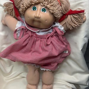 May include: A vintage Cabbage Patch Kids doll with a tan complexion, green eyes, and yarn hair styled in pigtails tied with red ribbons. The doll wears a red and white checkered dress with white lace trim.