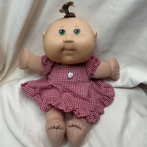 May include: A soft-bodied doll with a round face, green eyes, and a small tuft of brown hair tied at the top of the head. The doll wears a red and white gingham dress with ruffled sleeves, a ruffled hem, and a small white heart-shaped button.