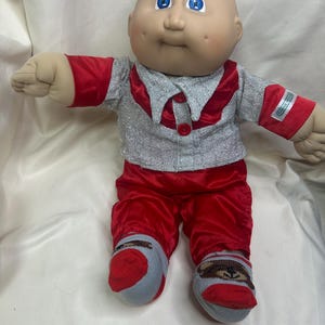 May include: A vintage Cabbage Patch Kid doll wearing a silver and red outfit. The doll has a bald head, blue eyes, and rosy cheeks. The outfit includes a silver jacket with red trim, red pants, and gray and red socks.