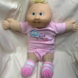 May include: A Cabbage Patch Kids doll with a bald head and green eyes. The doll wears a pink onesie with an owl graphic and pink and white striped socks. The doll's hair is a small tuft of yellow yarn.