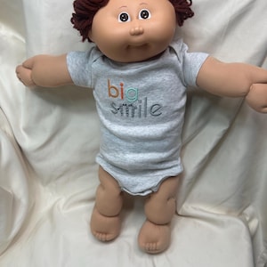 May include: A Cabbage Patch Kid doll with a light grey onesie. The onesie has the words "big smile" printed on the front in orange, teal, and grey. The doll has brown yarn hair and is posed with arms outstretched.