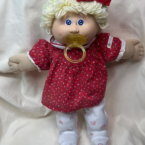 May include: A vintage Cabbage Patch Kid doll with blonde yarn hair and blue eyes. The doll wears a red floral print dress with white lace trim and white pants with rainbow accents. A red bow adorns the doll's hair, and it has a yellow pacifier.