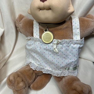 May include: A plush cat doll with a light brown body and a cream-colored face. The doll wears a white dress with colorful polka dots and a lace trim. It has blue eyes and a gold-colored medallion.