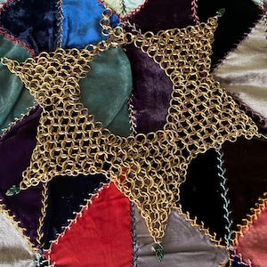 May include: A gold-toned chainmail collar necklace constructed from interlocking circular metal rings. The piece features a star-like geometric shape with pointed edges, each tipped with a small, dark green leaf-shaped charm accent.