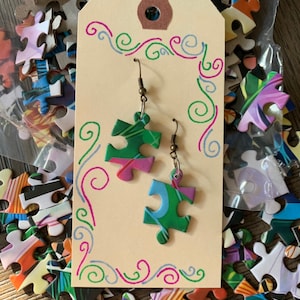 May include: A pair of puzzle piece earrings with a green, blue, and pink design. The earrings are hanging from a brown card with a white border and a decorative swirl design.