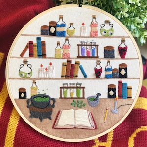 May include: Embroidered hoop art depicting a potion-making scene with shelves of books, bottles, and jars, a cauldron, a mortar and pestle, and an open book. The scene is stitched in various colours on a white background.