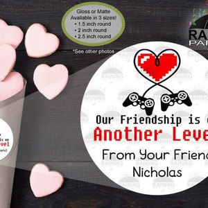 May include: A white round sticker with a red pixelated heart and two black pixelated game controllers connected by a cord. The text on the sticker reads "Our Friendship is on Another Level From Your Friend Nicholas".