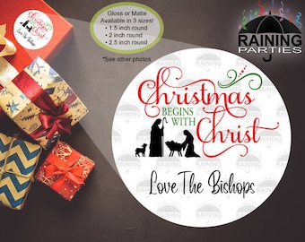 Pode incluir: Etiqueta redonda branca com o texto "Christmas Begins With Christ Love The Bishops" em letras vermelhas e pretas. A etiqueta também apresenta uma silhueta preta da cena da Natividade com Maria, José, o menino Jesus e um pastor.