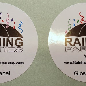 May include: Two round stickers with a black umbrella and colorful confetti. The text "RAINING PARTIES" is printed in black with the website "www.RainingParties.etsy.com". One sticker is labeled "Matte Label" and the other is labeled "Glossy Label".
