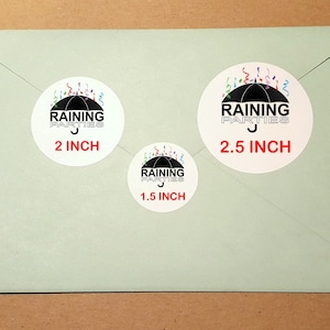 May include: Three round stickers with a black umbrella and colorful confetti design. The text "RAINING PARTIES" is on each sticker. The stickers are 1.5 inches, 2 inches, and 2.5 inches in diameter.