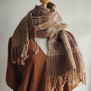 May include: A brown and beige scarf with vertical stripes and decorative fringe. The scarf is draped over a brown top on a mannequin. The scarf has a detailed pattern near the bottom edge.