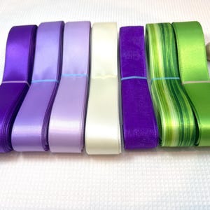 May include: A collection of colorful satin ribbons in shades of purple, lavender, cream, and green. The ribbons are neatly stacked and secured with thin bands. The arrangement includes solid and striped patterns, suitable for crafts and gift wrapping.