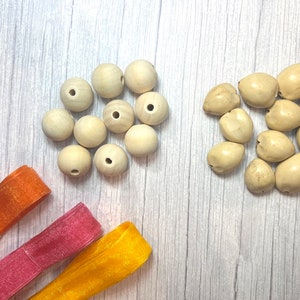 Peut inclure: Vue a&eacute;rienne de fournitures de bricolage sur une surface claire imitation bois. Deux groupes de perles sont pr&eacute;sents : des perles rondes en bois clair et des perles irr&eacute;guli&egrave;res blanc cass&eacute;. Des rubans bleus, oranges, roses et jaunes sont &eacute;galement visibles.