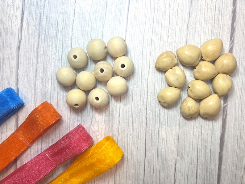 Peut inclure: Vue a&eacute;rienne de fournitures de bricolage sur une surface claire imitation bois. Deux groupes de perles sont pr&eacute;sents : des perles rondes en bois clair et des perles irr&eacute;guli&egrave;res blanc cass&eacute;. Des rubans bleus, oranges, roses et jaunes sont &eacute;galement visibles.