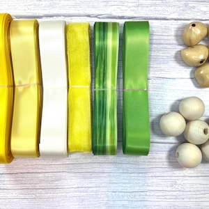 May include: Six spools of ribbon in shades of bright yellow, pale yellow, cream, mustard yellow, green striped, and solid green, accompanied by a collection of smooth, light-colored wooden beads and polished tan-colored oval beads.
