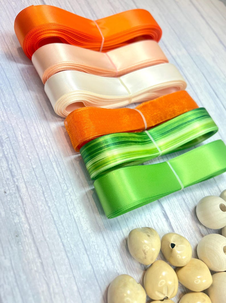 Peut inclure: Rubans de satin color&eacute;s, orange, p&ecirc;che, blanc et vert. Les rubans sont enroul&eacute;s et attach&eacute;s avec de la ficelle blanche. Des perles en bois sont &eacute;galement visibles, sugg&eacute;rant un th&egrave;me artisanal.