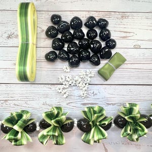 May include: Overhead shot of various craft supplies, including a roll of yellow and green striped ribbon, a bundle of black, polished stones, a small green fabric square, and a strand of white beads. A lei made of black stones, green and white leaves, and white beads is at the bottom.