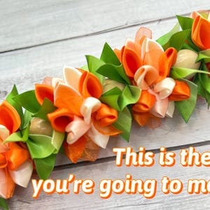 Peut inclure: Un lei fait &agrave; la main avec des fleurs en ruban orange, blanc et p&ecirc;che avec des feuilles vertes. Le texte "This is the lei you're going to make" est affich&eacute; en lettres orange.