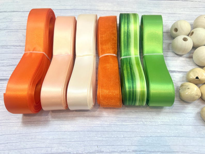 Peut inclure: Une collection de rubans color&eacute;s dans les tons orange, p&ecirc;che, cr&egrave;me et vert. Les rubans sont soigneusement regroup&eacute;s et dispos&eacute;s &agrave; c&ocirc;t&eacute; de perles en bois. Les rubans sont en satin et autres mat&eacute;riaux, parfaits pour l'artisanat et les d&eacute;corations.